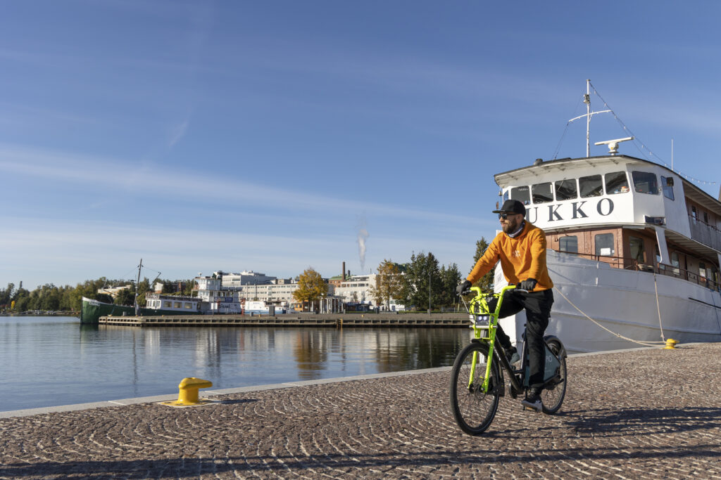 Ihminen pyöräilee kaupunkipyörällä Kuopion matkustajasatamassa. Taustalla näkyy Ravintolalaiva Ukko.