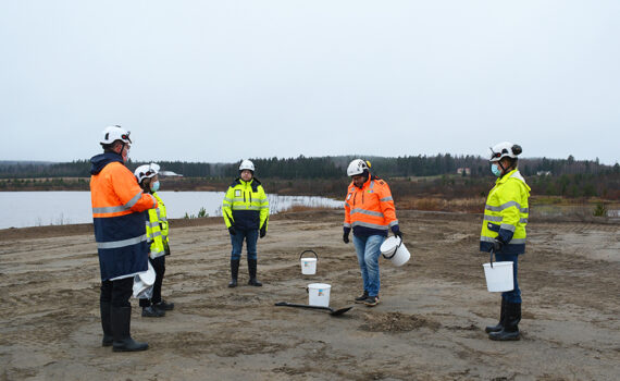 Viisi huomio- ja turva-asuista henkilöä seivovat ringissä hiekkakentällä. Osalla ämpärit kädessa. Lapio maassa. Taustalla näkyy Keltin allas.