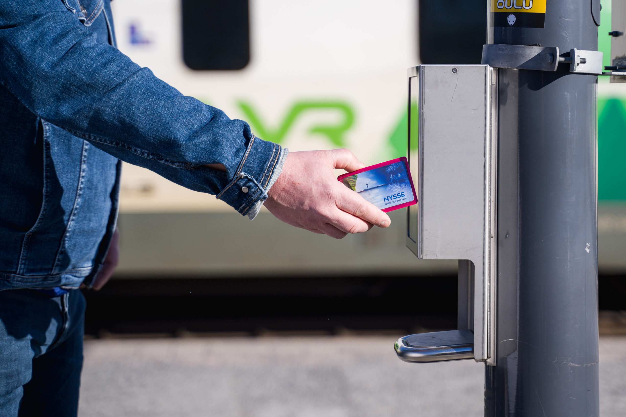 Kuvassa Nyssen matkakorttia leimataan juna-asemalla ennen junaan nousua. Kortin ja leimauslaitteen takana näkyy VR:n juna.