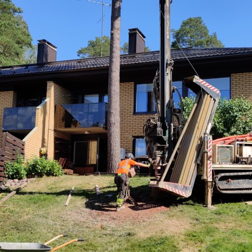 Rakentaja poraa maalämpökaivoa rinnetontilla. Taustalla keltatiilinen kaksikerroksinen paritalo.