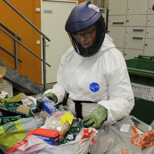 Kuvassa henkilö tekee sekajätteen koostumustutkimusta suojavarusteissa. Hänen edessään pöydällä on sekajätteitä lajiteltavana.