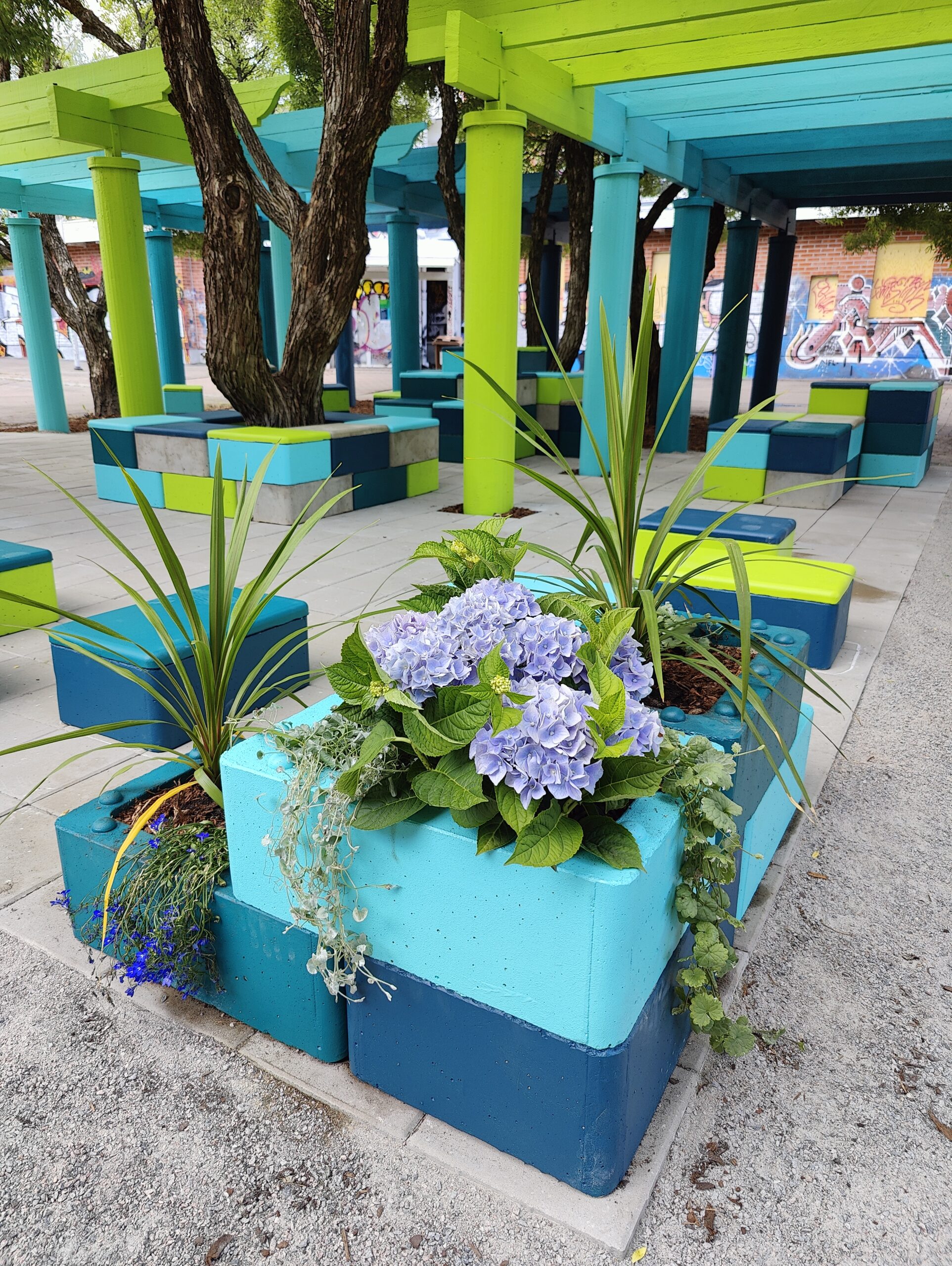 Kuvassa sinisävyisiä Cubeco-istutuslaatikoita, joissa erilaisia kasvi- ja kukkaistutuksia. Taustalla sinivihreät pergolat puiden suojassa. 