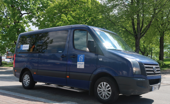Ukikyyti: tumman sininen Wolkswagen tila-auto kyljen puolelta kuvattuna. Taustalla vihreitä puita.