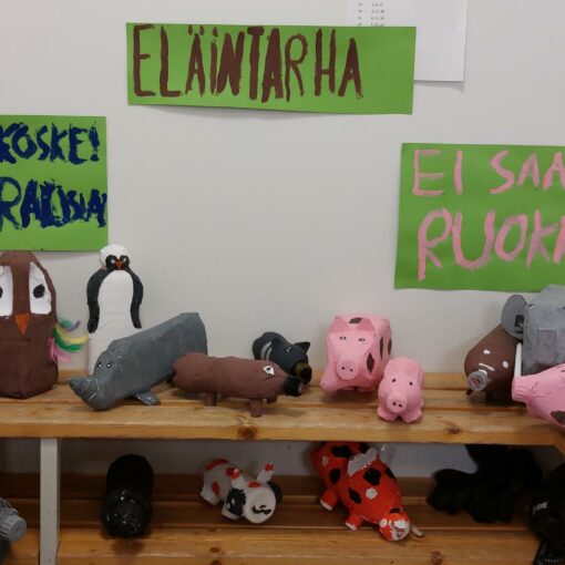 Kuvassa koululaisten tekemiä eläinhahmoja puupenkeillä, taustalla seinällä kolme vihreää kylttiä, joihin on kirjoitettu Eläintarha, Ei saa ruokkia, ja Älä koske! - vaarallisia