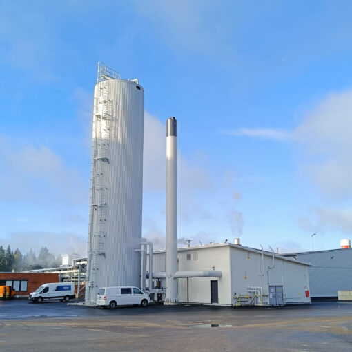 Advenin energialaitos lämpöakkuineen Säkylässä Apetitin pakastetehtaan vieressä. Kuvassa energialaitos ja lämpöakku, jotka ovat valkoinen matala rakennus ja sen vieressä korkea valkoinen torni. Laitoksen pihaan on parkkeerattu kaksi pakettiautoa. Taustalla sininen taivas ja muutama pilviharso.