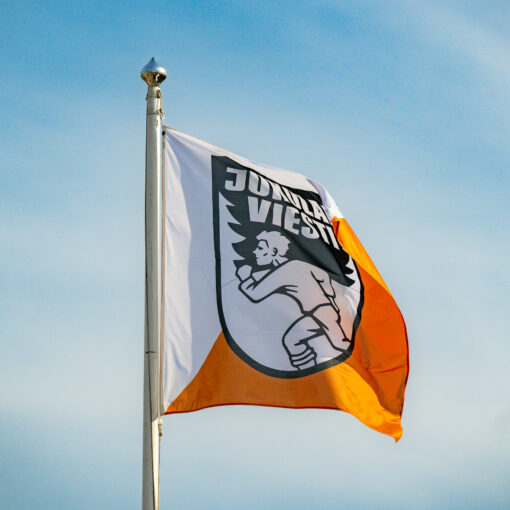 Jukolan viestin lippu salossa. Lipussa puolet oranssia ja puolet valkoista. Logossa animoitu mies juoksee. Taustalla sininen taivas ja harsopilviä.