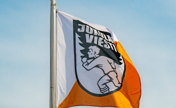 Jukolan viestin lippu salossa. Lipussa puolet oranssia ja puolet valkoista. Logossa animoitu mies juoksee. Taustalla sininen taivas ja harsopilviä.