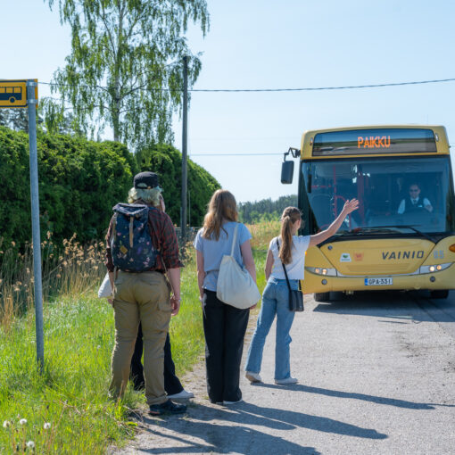 Kesäisen maantien varressa bussipysäkki. Pysäkillä seisoo neljä henkilöä, joista ensimmäinen viittaa keltaista bussia pysähtymään. Bussin keulassa lukee "PAIKKU".