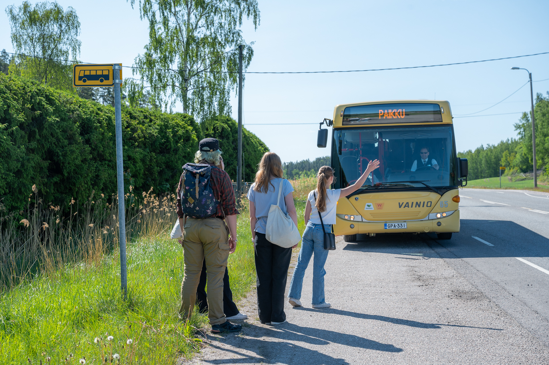 Kesäisen maantien varressa bussipysäkki. Pysäkillä seisoo neljä henkilöä, joista ensimmäinen viittaa keltaista bussia pysähtymään. Bussin keulassa lukee "PAIKKU".