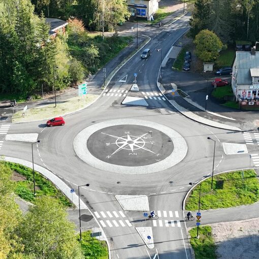 Sipoon Söderkullan liikenneympyrä lintuperspektiivistä. Ympyrään on ajamassa yksi auto ja siitä on poistumassa toinen. Liikenneympyrän keskellä on maaluttuna kompassi. Reunalla vanha rakennus. Taustalla vihreää nurmea ja puita.