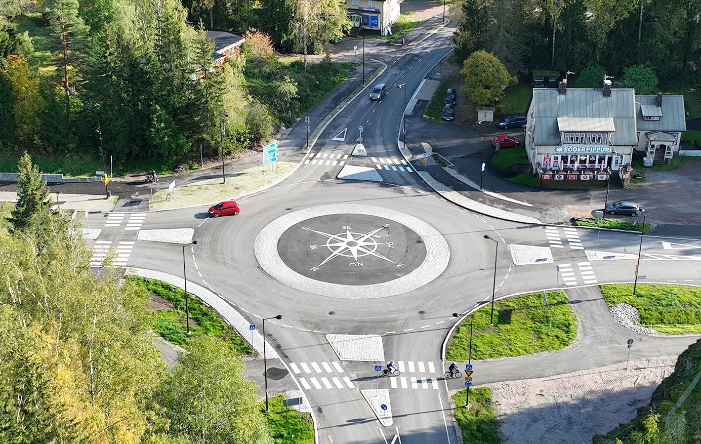 Sipoon Söderkullan liikenneympyrä lintuperspektiivistä. Ympyrään on ajamassa yksi auto ja siitä on poistumassa toinen. Liikenneympyrän keskellä on maaluttuna kompassi. Reunalla vanha rakennus. Taustalla vihreää nurmea ja puita.
