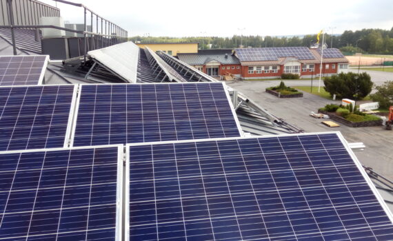 Aurinkopaneeleja, taustalla punainen rakennus ja peltomaisema.