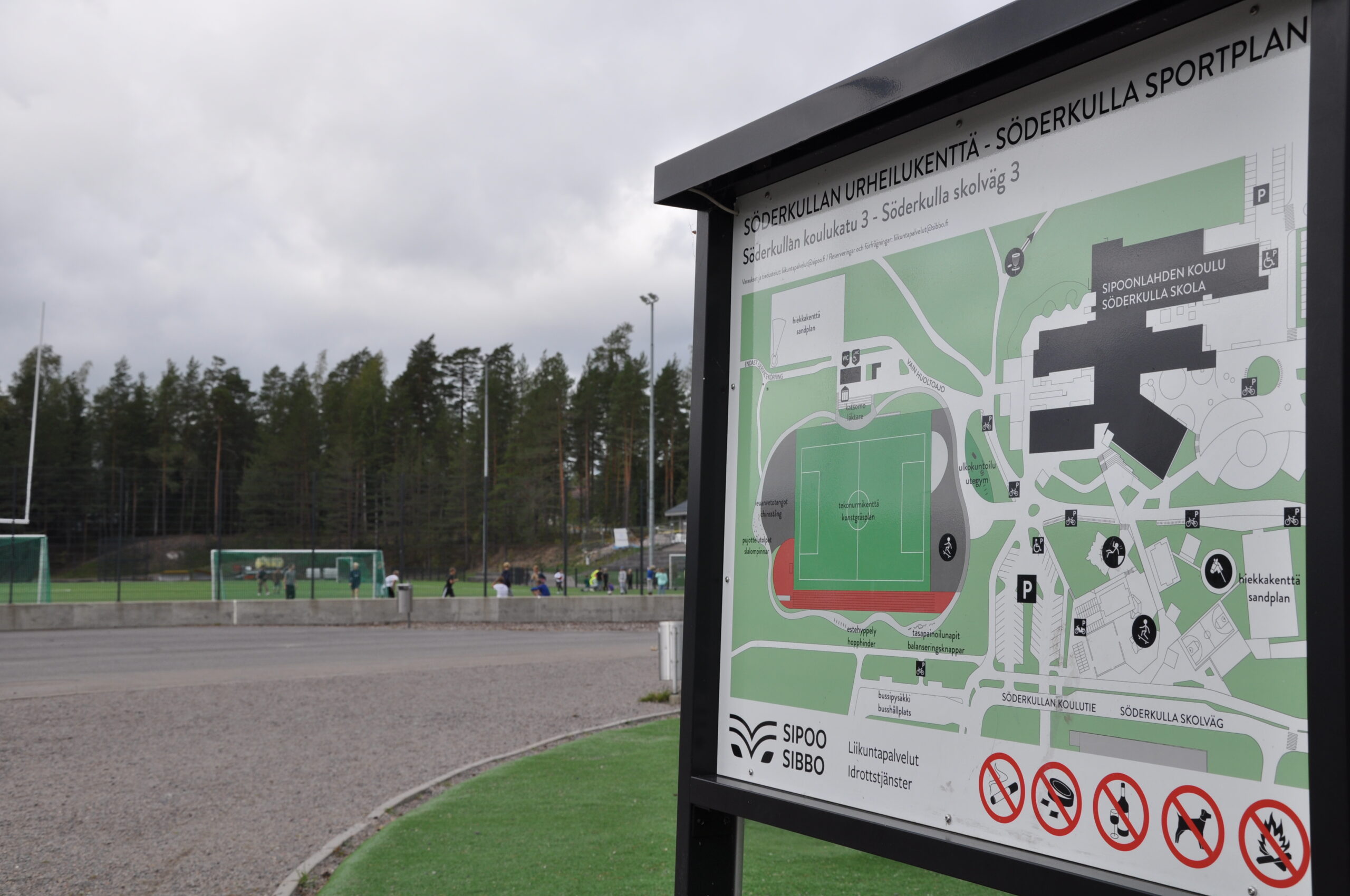 Oikealla Söderkullan urheilukentän infotaulu. Taustalla hiekkarata ja tekonurmikenttä jalkapallokenttineen sekä havupuita.