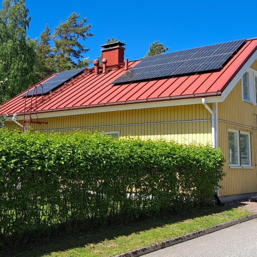 Keltainen puutalo, jonka punaisella peltikatolla on asennettuna aurinkopaneelit. Talon edustalla on vihreä pensasaita. Sää on aurinkoinen ja taivas kirkkaan sininen.