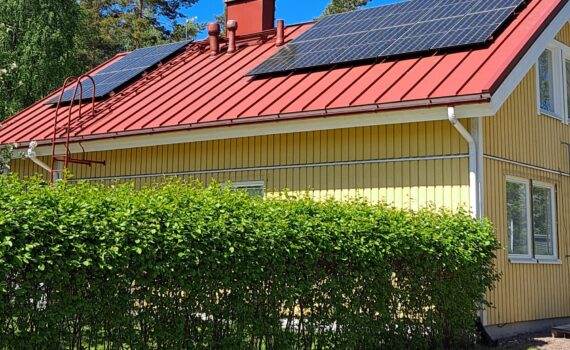 Keltainen puutalo, jonka punaisella peltikatolla on asennettuna aurinkopaneelit. Talon edustalla on vihreä pensasaita. Sää on aurinkoinen ja taivas kirkkaan sininen.