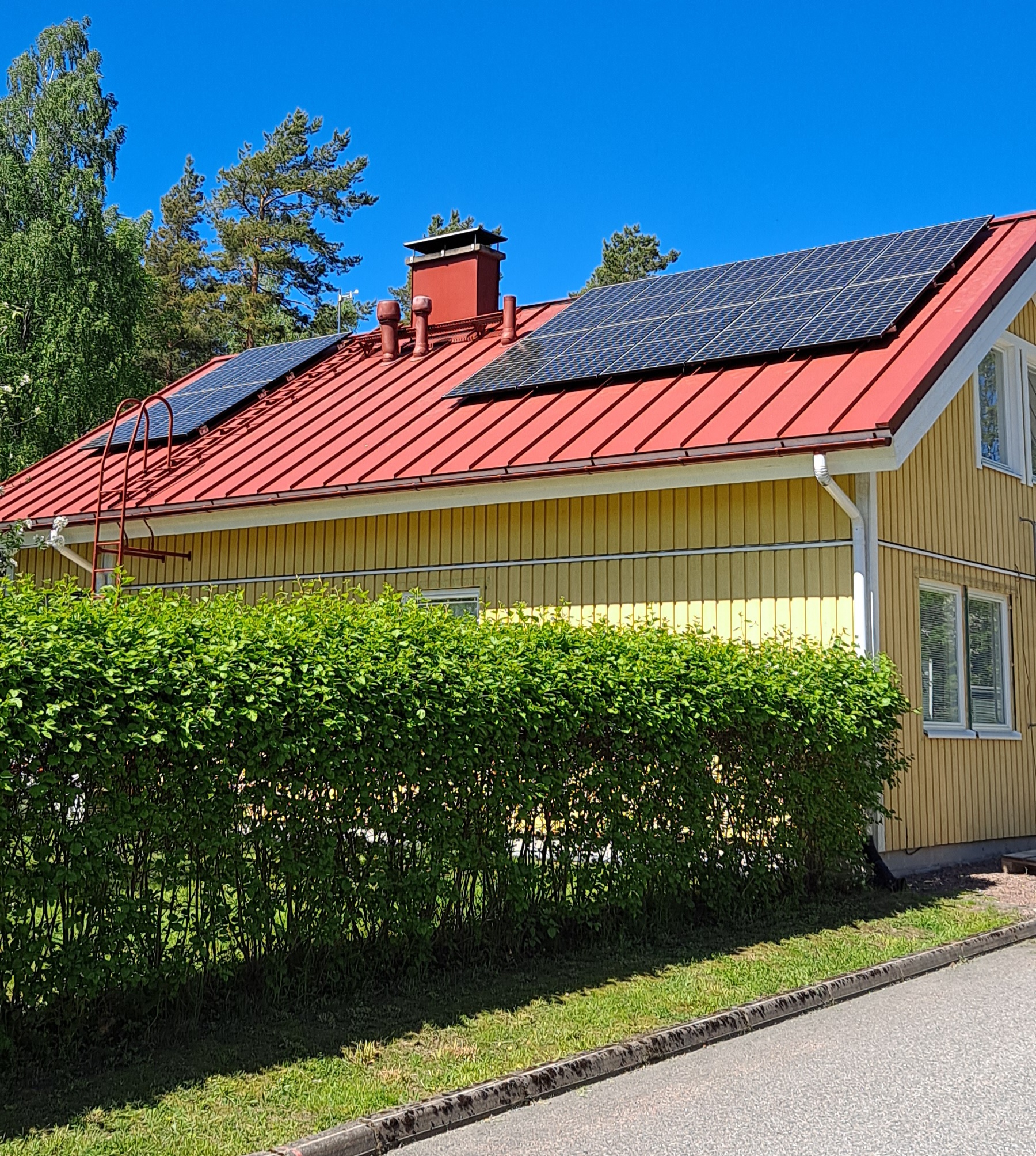 Keltainen puutalo, jonka punaisella peltikatolla on asennettuna aurinkopaneelit. Talon edustalla on vihreä pensasaita. Sää on aurinkoinen ja taivas kirkkaan sininen.