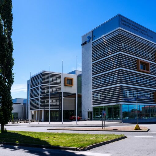 SAMKin Porin kampus. Kulmikas moderni rakennus. Etualalla asfalttia, nurmikaistale ja kaksi havupuuta. Taivas on sininen ja kirkas.