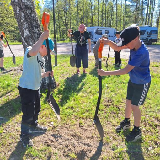 Etualalla kaksi poikaa iskemässä lapioita maahan. Taustalla muita henkilöitä myös lapiot käsissään. Henkilöt ovat poistamassa aiempaa kasvillisuutta niityn tieltä.