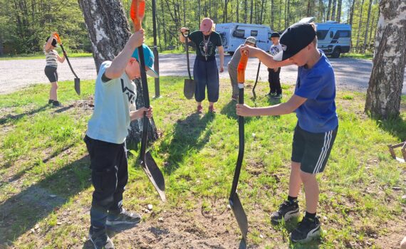 Etualalla kaksi poikaa iskemässä lapioita maahan. Taustalla muita henkilöitä myös lapiot käsissään. Henkilöt ovat poistamassa aiempaa kasvillisuutta niityn tieltä.
