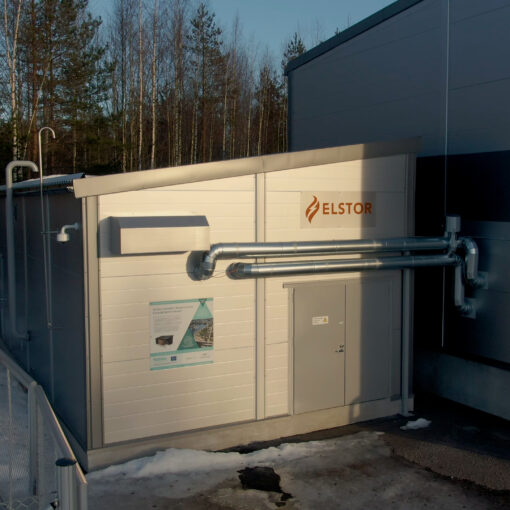 Lämpöakkurakennus isomman rakennuksen kyljessä. Akun seinässä Elstorin kyltti ja Canemure-hankkeen infokyltti sekä isot metalliset putket. Alue on rajattu aidoin. Maassa on lunta.
