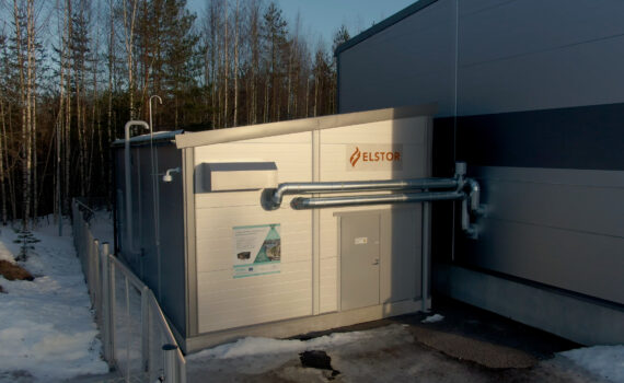 Lämpöakkurakennus isomman rakennuksen kyljessä. Akun seinässä Elstorin kyltti ja Canemure-hankkeen infokyltti sekä isot metalliset putket. Alue on rajattu aidoin. Maassa on lunta.