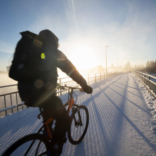 Pyöräilijä Oulussa talvipakkasella. Pyöräilijällä on selässään iso laukku. Taivas on sininen, aurinko paistaa ja maassa on lunta.