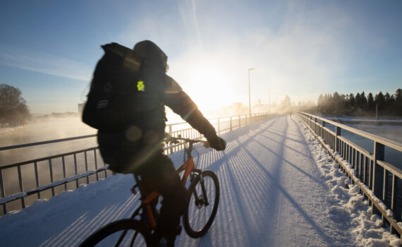 Pyöräilijä Oulussa talvipakkasella. Pyöräilijällä on selässään iso laukku. Taivas on sininen, aurinko paistaa ja maassa on lunta.