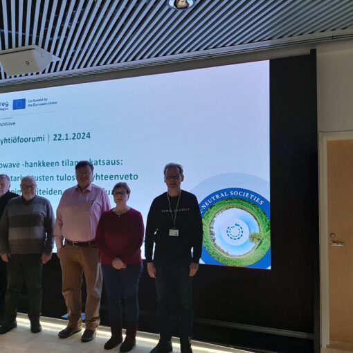 Kuusi taloyhtiöfoorumin kokouksen osallistujaa seisoo valkokankaan edessä. Valkokankaalla olevassa esityksessä lukee "Taloyhtiöfoorumi, 22.1.2024. Renowave-hankkeen tilannekatsaus".