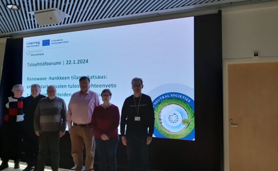 Kuusi taloyhtiöfoorumin kokouksen osallistujaa seisoo valkokankaan edessä. Valkokankaalla olevassa esityksessä lukee "Taloyhtiöfoorumi, 22.1.2024. Renowave-hankkeen tilannekatsaus".