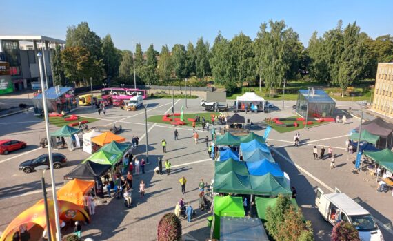 Näytteillepanopisteiden telttoja ja ständejä parkkipaikalla lintuperspektiivistä kuvattuna. Pisteillä kiertää ihmisiä. Taivas on sininen ja taustalla näkyy puita.