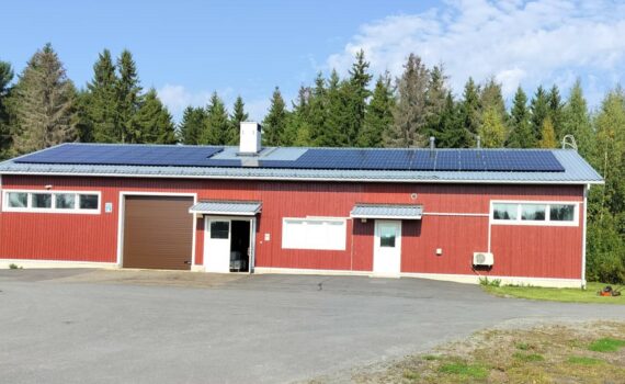 Rääkkylän jätevedenpuhdistamo. Punainen lautarakennus, jonka katolla on aurinkopaneelit. Edessä asfalttikenttä, sivulla nurmea ja ruohonleikkuri, taustalla metsää.