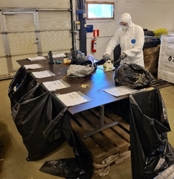 Suojapuvulla varustautunut työntekijä lajittelee jätteitä pöydän ääressä mustiin jätesäkkeihin.