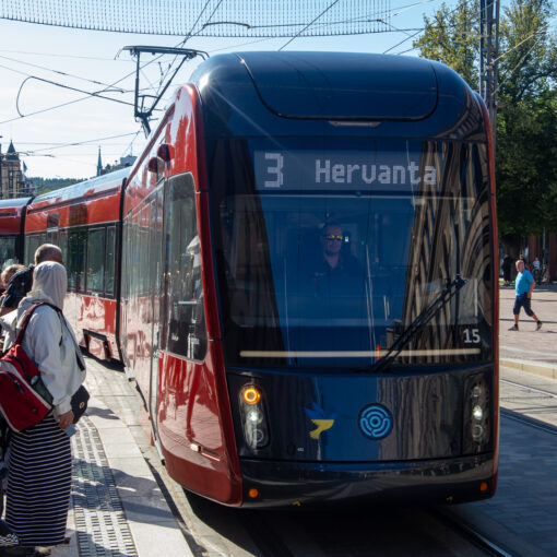 Punainen raitiovaunu edestä kuvattuna. Raitiovaunun ikkunassa lukee "3 Hervanta". Aurinkoinen kesäpäivä. Ihmiset odottavat raitiovaunua pysäkillä.