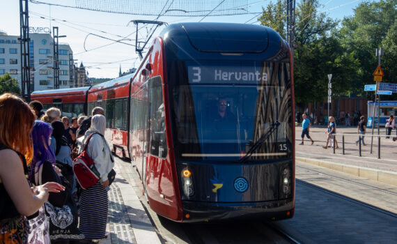 Punainen raitiovaunu edestä kuvattuna. Raitiovaunun ikkunassa lukee "3 Hervanta". Aurinkoinen kesäpäivä. Ihmiset odottavat raitiovaunua pysäkillä.