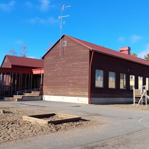 Ruskea hirsirakennus, sininen taivas, vieressä leikkipuisto ja asfalttikatu.