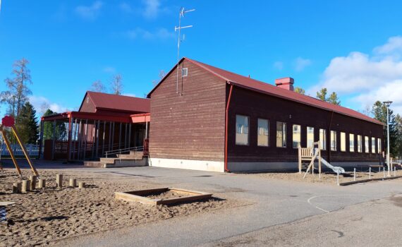 Ruskea hirsirakennus, sininen taivas, vieressä leikkipuisto ja asfalttikatu.