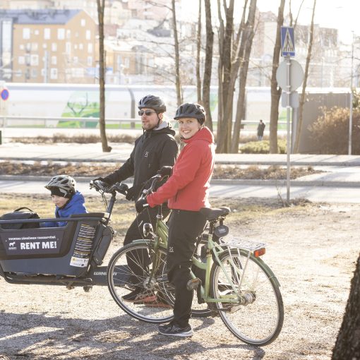 Pyöräilijät pysähtyneenä kadun varteen. Etualan pyäräilijällä on päällään punainen takki. Takana oleva pyöräilijä on pukeutunut mustiin ja hänen pyörässään on keulassa lapsenkuljetuskaukalo.