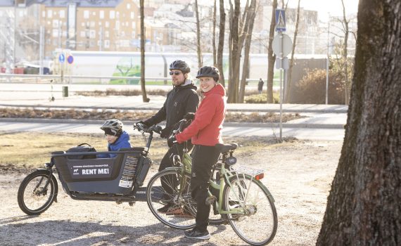 Pyöräilijät pysähtyneenä kadun varteen. Etualan pyäräilijällä on päällään punainen takki. Takana oleva pyöräilijä on pukeutunut mustiin ja hänen pyörässään on keulassa lapsenkuljetuskaukalo.